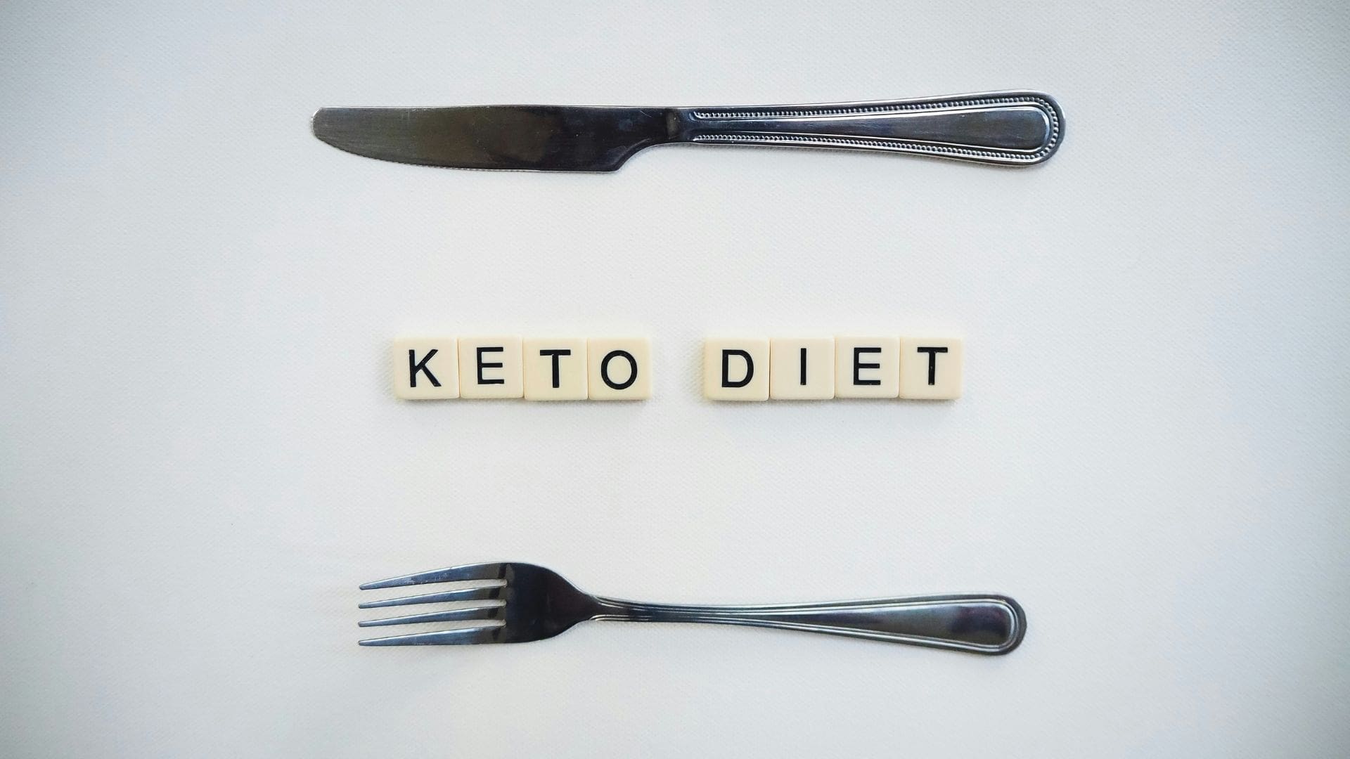 low carb MAIN scrabble tiles spelling out keto diet on white table sitting in between silver knife and fork