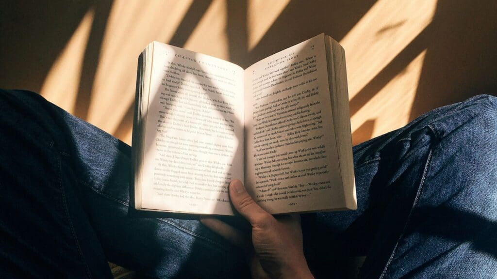 POV of man holding book in his hand while sitting cross-legged in a pair of jeans