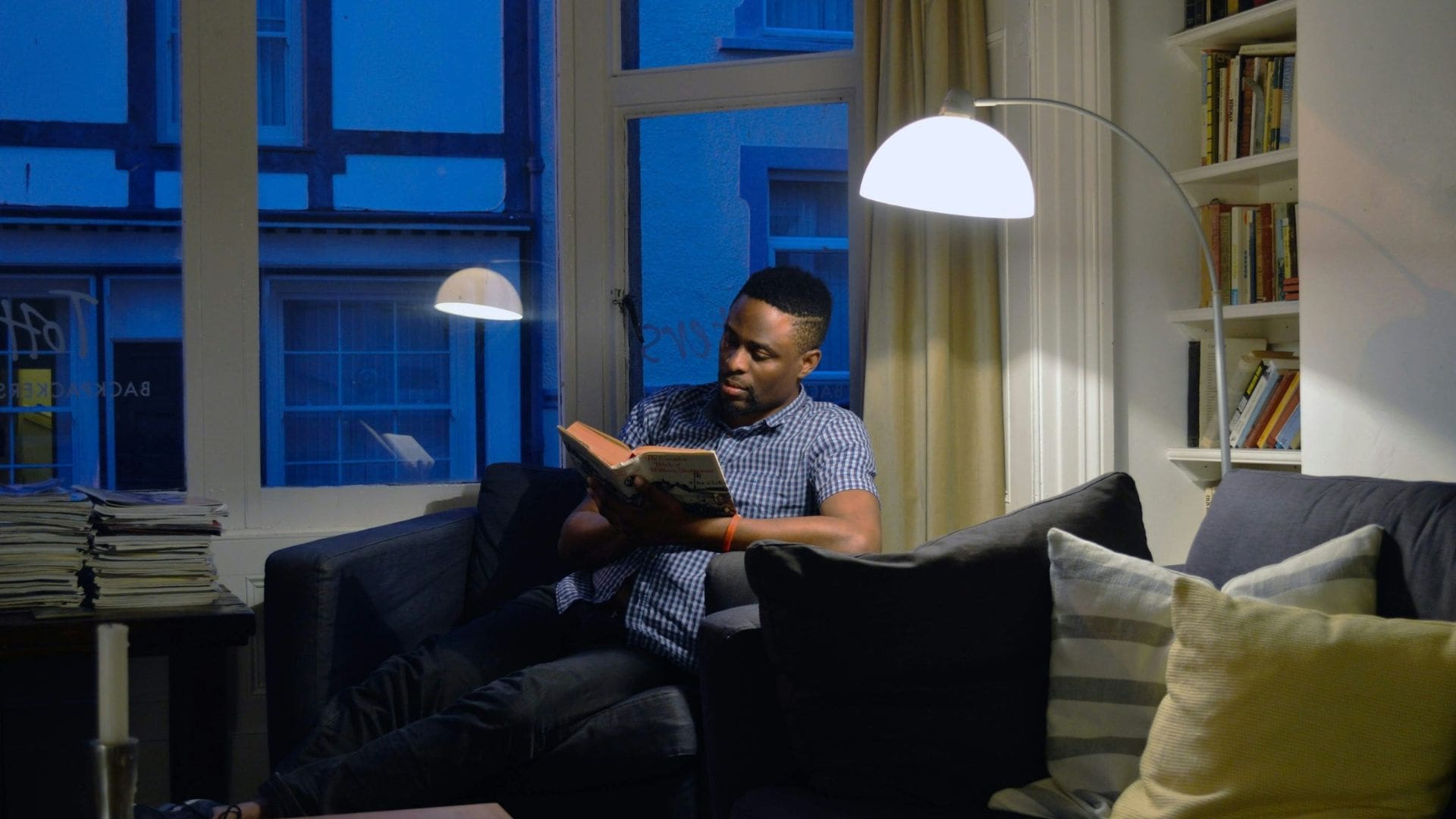 man sitting in a dimly lit lounge area reading a book under a standing light